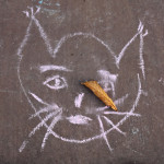 Naive children drawing of a cat, made with pink chalk on the sidewalk. The simple animal trace has some human features such as female eyes and mouth. On top of the face, near the whiskers there's a dried out autumn leaf.